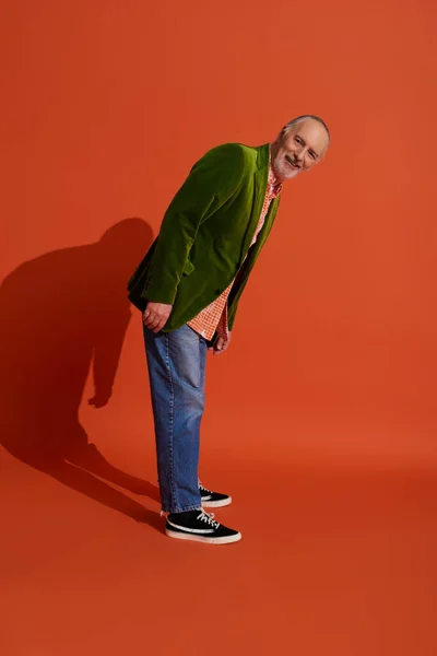 stock image full length of joyful and bearded senior man in trendy casual clothes standing and looking at camera on red orange background, green velour blazer, blue denim jeans, happy aging concept