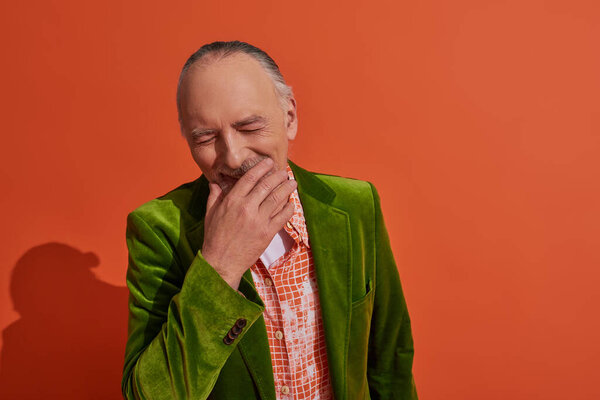 happy aging, overjoyed senior man covering mouth with hand and laughing with closed eyes on red orange background, green velour blazer, trendy shirt, fashion and age concept