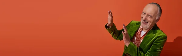 stock image happy and trendy aging concept, excited grey haired and bearded senior man in green velour blazer showing stop gesture and laughing with closed eyes on red orange background, banner with copy space