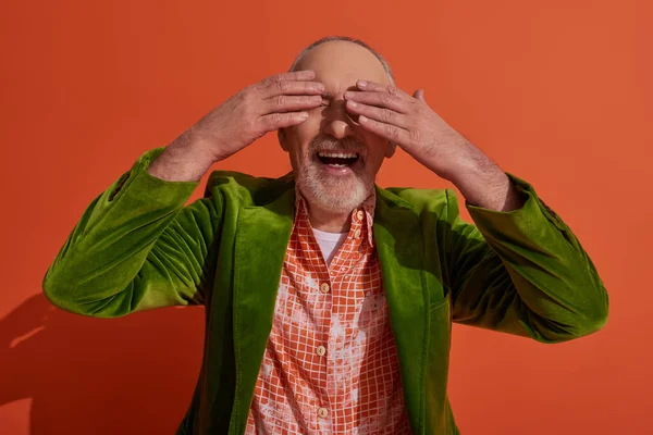 stock image happy aging lifestyle, excited senior man smiling and covering eyes with hands while waiting for surprise on red orange background, green velour blazer, trendy shirt, fashion and style concept
