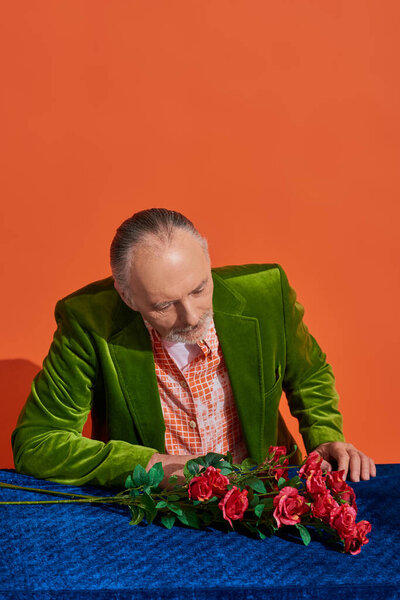 melancholy, memories, thoughtful senior man in green and fashionable velvet blazer looking at fresh red roses on blue velour cloth while sitting on vibrant orange background with copy space