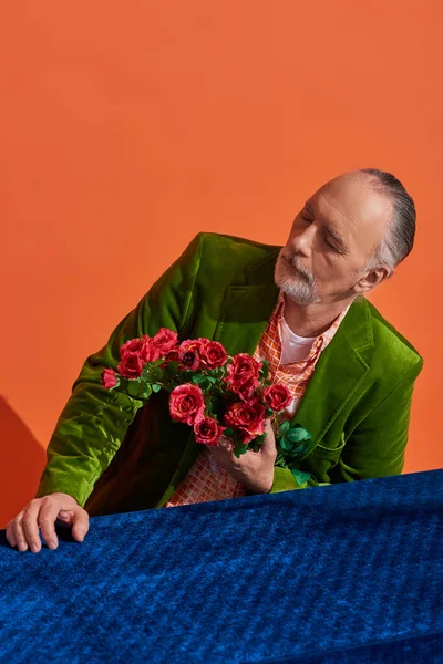 stock image memories, melancholy, bearded senior man in green velvet blazer holding bouquet of red roses while sitting at table with blue velour cloth on vibrant orange background, aging population concept