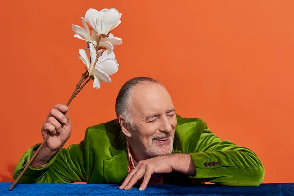 stock image cheerful and charismatic senior man with closed eyes holding white orchid flower while sitting at table with blue velour cloth on vibrant orange background, green velour blazer, personal style