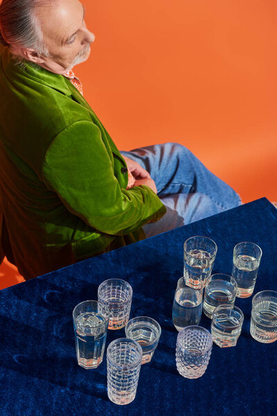 stylish and thoughtful senior man in green velvet blazer sitting near glasses with fresh pure water on table with blue velour cloth, symbolism, life fullness concept, high angle view