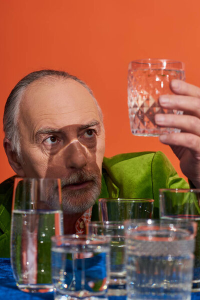 senior and thoughtful grey haired man in green velvet blazer looking at glass full of pure water near table with blue cloth on orange background, aging population, symbolism, life fullness concept