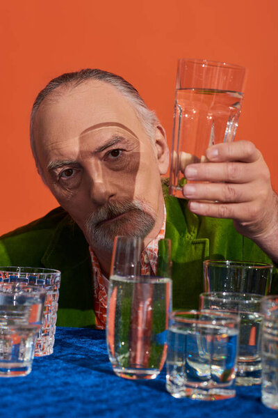 senior bearded man in fashionable and green velvet blazer holding glass of clear water and looking at camera on orange background, aging population, symbolism, life fullness concept
