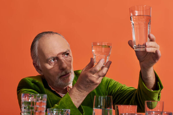 thoughtful man with grey hair and beard, wearing green velvet blazer, looking at glasses with pure water on orange background, aging population, symbolism, life fullness concept 