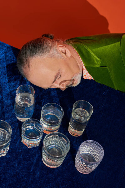 high angle view or aged grey haired man with closed eyes laying on table with blue velour cloth and glasses with pure water on orange background, aging population, symbolism, life fullness concept