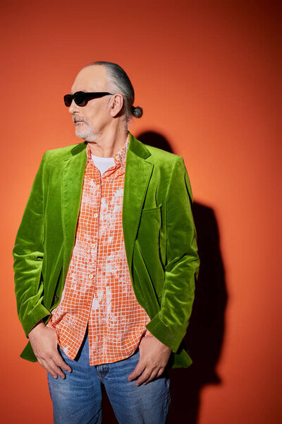 старший бородатый мужчина в темных солнцезащитных очках, модная рубашка и зеленый велюровый блейзер стоят с большими пальцами в карманах синих джинсов и смотрят в сторону на красный и оранжевый фон с тенью