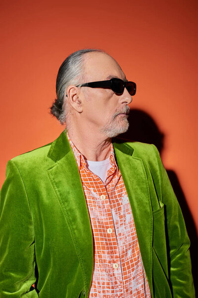 fashion look, senior male model with grey hair and beard looking away on red and orange background with shadow, dark sunglasses, trendy shirt, green velour blazer, stylish lifestyle of aging man