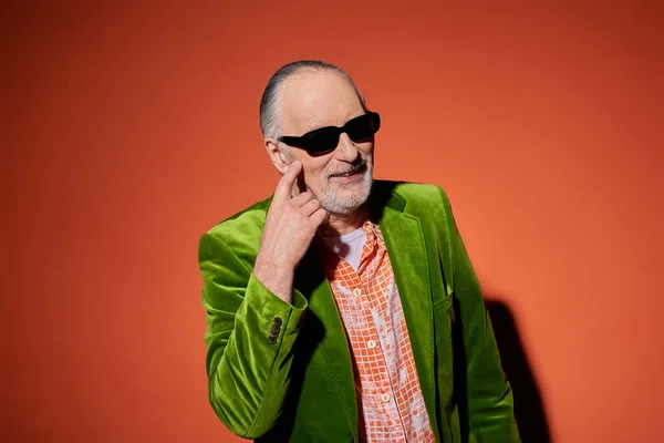 stock image playful and fashionable senior man pointing at cheek while asking for kiss on red and orange background, trendy shirt, green velour blazer, dark sunglasses, vibrant aging personality concept
