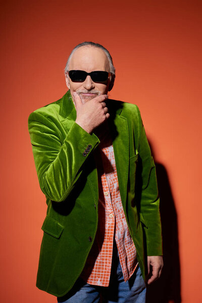 smiling senior man in dark sunglasses and green velour blazer touching beard and looking at camera on red and orange background, personal style, happy aging lifestyle concept
