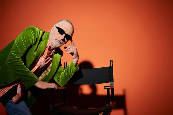stock image thoughtful aged man in dark sunglasses and green velour blazer posing with chair and looking at camera on red and orange background with shadow, fashion look, stylish lifestyle