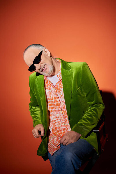 joyful senior man with grey hair and beard sitting on chair and smiling at camera on red and orange background, dark sunglasses, green velour blazer, fashion trend, happy aging concept