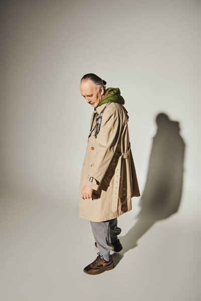 full length of grey haired and bearded senior man in beige trench coat and green hoodie looking down while standing on grey background with shadow, stylish casual wear, fashionable aging concept