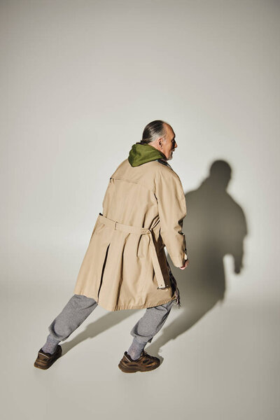 full length of elderly man in beige trench coat and green hoodie standing in expressive pose on grey background with shadow, casual fashion, stylish and positive aging concept