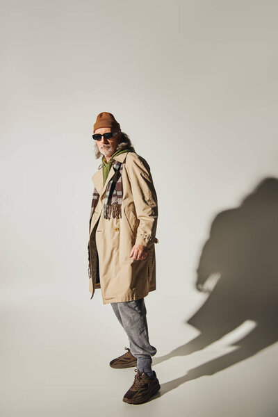 full length of fashionable senior model in dark sunglasses, beanie hat, beige trench coat and plaid scarf standing on grey background with shadow, positive and trendy aging concept