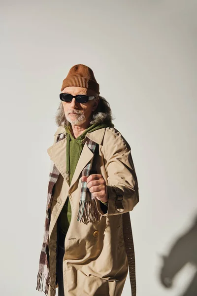 stock image confident senior man in dark sunglasses and hipster style attire standing with clenched fist and looking at camera on grey background, beanie hat, beige trench coat, fashionable aging concept