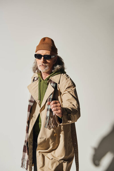 confident senior man in dark sunglasses and hipster style attire standing with clenched fist and looking at camera on grey background, beanie hat, beige trench coat, fashionable aging concept