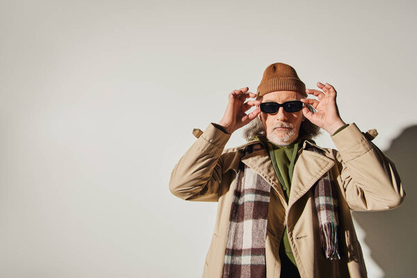 bearded senior man in beanie hat, plaid scarf and stylish trench coat looking at camera while standing on grey background, hipster style, expressive individuality, trendy aging concept