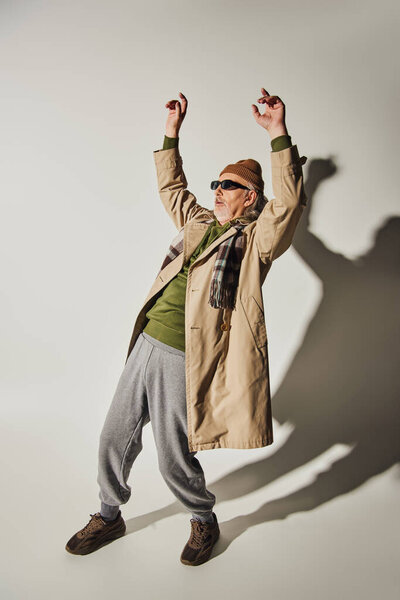 full length of expressive hipster style senior man in dark sunglasses, beanie hat, beige trench coat and plaid scarf posing with raised hands on grey background with shadows, dance