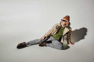 full length of fashionable senior male model sitting and looking away on grey background with shadow, dark sunglasses, beanie hat, beige trench coat, hipster style, positive aging concept clipart