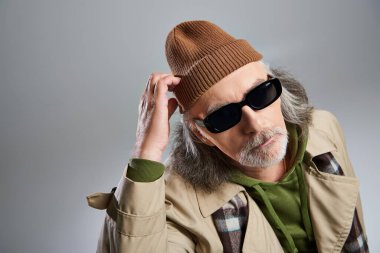 Koyu güneş gözlüklü ve bej renkli trençkotlu bir adamın portresi. Şapkaya dokunuyor ve gri arka plandaki kameraya bakıyor. Hipster modası, şık ve pozitif yaşlanma kavramı.