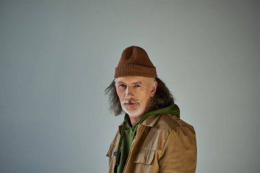 Gri saçlı, sakallı, bereli ve kahverengi ceketli yaşlı adam gri arka planda dururken kameraya bakıyor, hipster tarzı model, yaşlanan nüfus yaşam tarzı konsepti