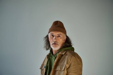 Düşünceli ve yılgın hipster tarzı şapka ve kahverengi ceketli son sınıf öğrencisi gri arka planda, yaşlanan nüfus yaşam tarzı konseptinde kameraya bakıyor.