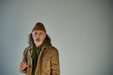 Gri saçlı, sakallı, bereli ve kahverengi ceketli yaşlı bir adam gri arka planda kameraya bakıyor, hipster tarzı, yaşlanan nüfus yaşam tarzı konsepti