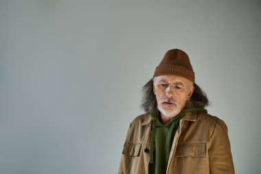 Gri saçlı, sakallı, bereli ve kahverengi ceketli yaşlı adam gri arka planda duruyor, hipster tarzı, etkileyici kişilik, yaşlanan nüfus yaşam tarzı konsepti