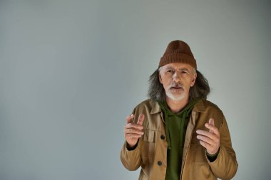 Üzgün ve endişeli, şapkalı, kahverengi ceketli yaşlı sakallı adam gri arka planda kameraya bakıyor, hippi kıyafetleri, yaşlanan nüfus yaşam tarzı konsepti.