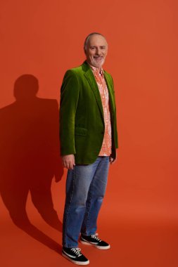 Tam boy iyimser yaşlı erkek model kırmızı turuncu arka planda duruyor ve kameraya gülümsüyor, modaya uygun gömlek, yeşil kadife ceket, mavi kot pantolon, günlük kıyafet, mutlu yaşlanma konsepti