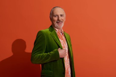 senior grey haired man, charismatic and cheerful, posing in trendy shirt and green velour blazer on red orange background, looking at camera, smiling, positive and fashionable aging concept clipart