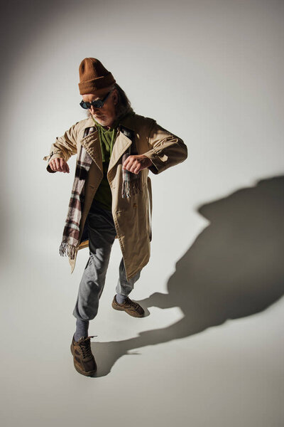 aging with style concept, full length of senior hipster man in dark sunglasses, beanie hat, beige trench coat and plaid scarf standing in stylish pose on grey background with shadow