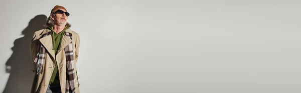 流行のカジュアルな服と影と離れて見て灰色の背景に立って暗いサングラスで高齢者 ヒップスタースタイル ビーニー帽子 ベージュトレンチコート 再生スカーフ コピースペースとバナー — ストック写真