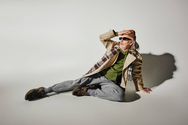 expressive personality, hipster style, full length of senior man in dark sunglasses and trendy casual attire touching beanie hat and looking away while sitting on grey background with shadow