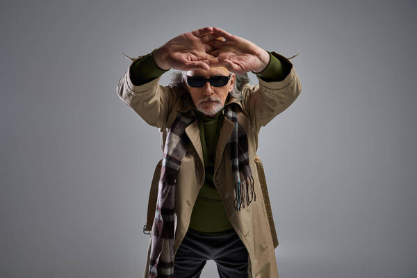 fashion shoot of senior hipster man in fashionable casual clothes and dark sunglasses standing with clenched stretched hands and looking at camera on grey background, fashion and age concept