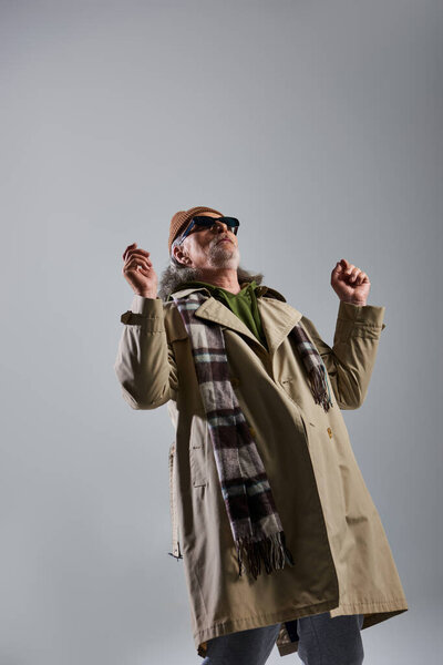 выразительная личность, старший и бородатый хипстер мужчина в темных солнцезащитных очках и стильной повседневной одежде глядя вверх, стоя на сером фоне, модная и позитивная концепция образа жизни