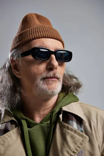 stock image portrait of confident grey haired and bearded senior man in beanie hat, dark sunglasses and beige trench coat looking away on grey background, hipster style, fashion and age concept