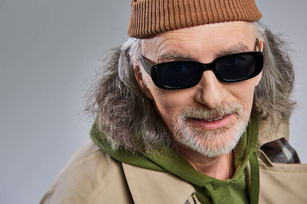 portrait of smiling senior man with grey hair and groomed beard, in dark sunglasses, beanie hat and trench coat looking at camera on grey background, hipster fashion, happy and trendy aging concept