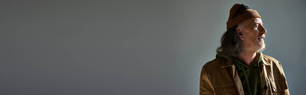 delighted hipster style senior man in beanie hat and brown jacket standing and smiling with closed eyes on grey background, happy and fashionable aging concept, banner with copy space