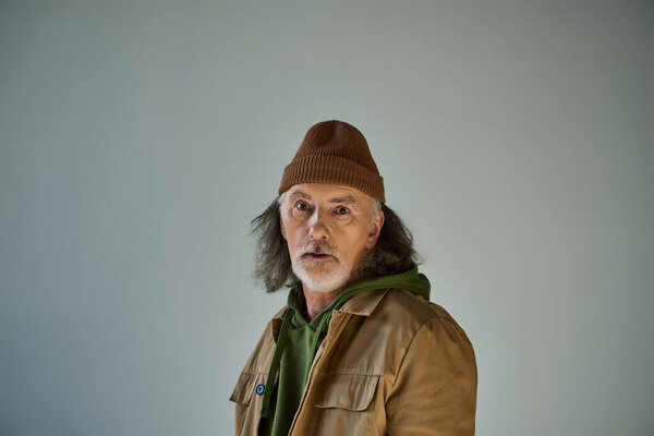 thoughtful and discouraged hipster style senior man in beanie hat and brown jacket looking at camera while standing on grey background, aging population lifestyle concept
