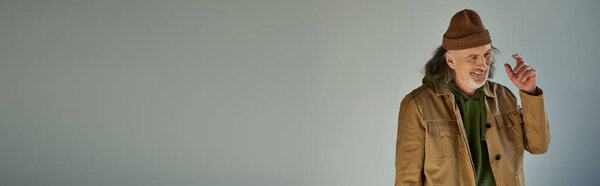 cheerful and bearded senior man gesturing and smiling on grey background, beanie hat, brown jacket, hipster style, happy and fashionable aging concept, banner with copy space