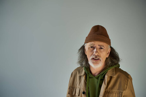 расстроен старший мужчина с оскорбленным выражением лица глядя на камеру на сером фоне, седые волосы, бороду, шапку шапки, коричневую куртку, хипстерскую моду, старение населения концепции образа жизни