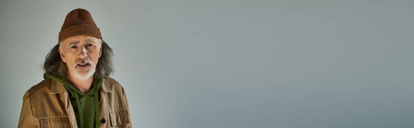 frustrated senior man with worried face expression standing on grey background, grey hair, brown jacket, beanie hat, hipster fashion, aging population lifestyle concept, banner with copy space