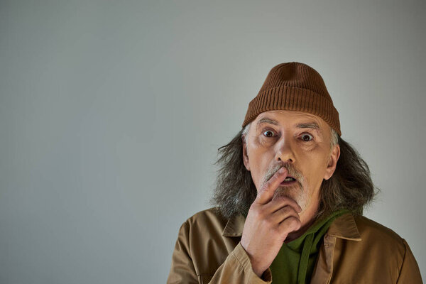 impressed and shocked senior man with grey hair and beard touching lips and looking at camera on grey background, beanie hat, brown jacket, hipster fashion, aging population lifestyle concept