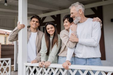 parents day, middle aged parents hugging and looking away near teenage daughter and adult son on porch of summer house, family celebration, bonding, modern parenting, translation of tattoo: harm none clipart