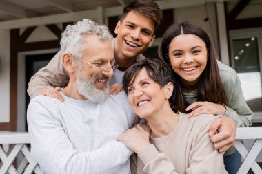 Ebeveynler günü, orta yaşlı ebeveynler genç kız ve genç yetişkin oğullarıyla birlikte yazlık evin verandasında, aile toplantısı, bağ kurma, modern ebeveynlik, hatırlanacak anlar. 