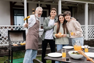 Aile fotoğrafı, mutlu ebeveynler günü, neşeli orta yaşlı ebeveynler genç kız ve genç yetişkin oğluyla bbq partisi, heyecanlı baba ızgara mısırla maşa tutuyor, kameraya bakıyor,